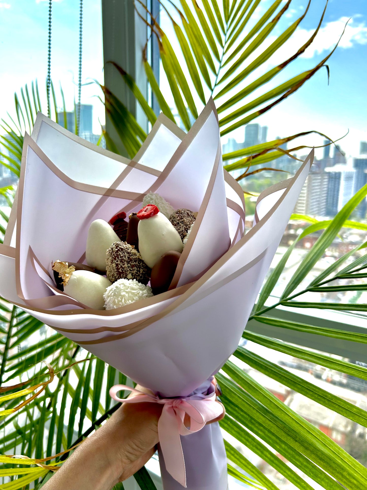 hand holds bouquet of chocolate covered strawberries in purple wrapping, green leafs on the background