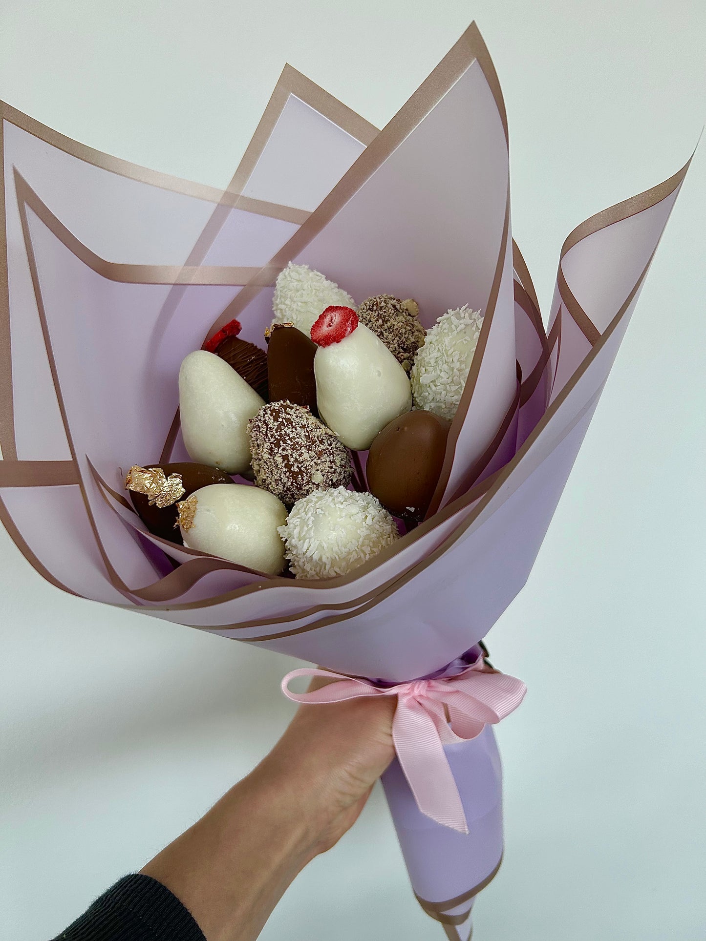 hand holds bouquet of chocolate covered strawberries in purple wrapping on white background