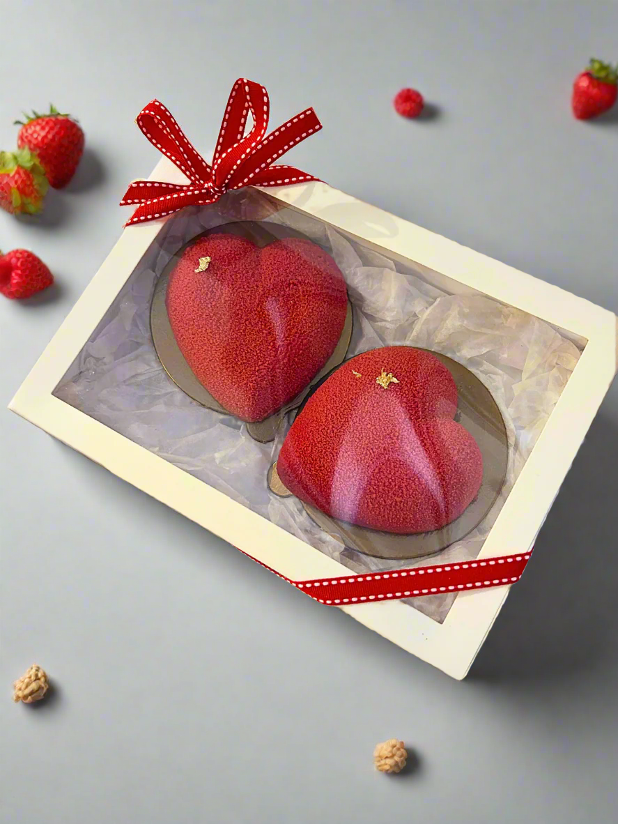 two mini heart shaped mousse dessert in white box on the grey background with some strawberries 